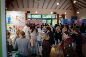 Auf dem Bild ist eine Messehalle zu sehen. Viele Menschen unterschiedlichen Alters unterhalten sich angeregt mit Aussteller und Ausstellerinnen über Ausbildungs- und Karrieremöglichkeiten.